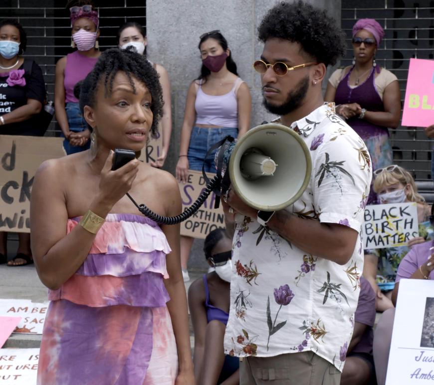 At a sidewalk rally in New York City, a black woman in a purple dress speaks into a megaphone. Next to her is a younger black man with sunglasses, a moustache, a beard, and a tropical shirt. Men and women -- many of them black but a few white and Asian -- stand and sit behind them, holding signs with slogans such as "Black Birth Matters."