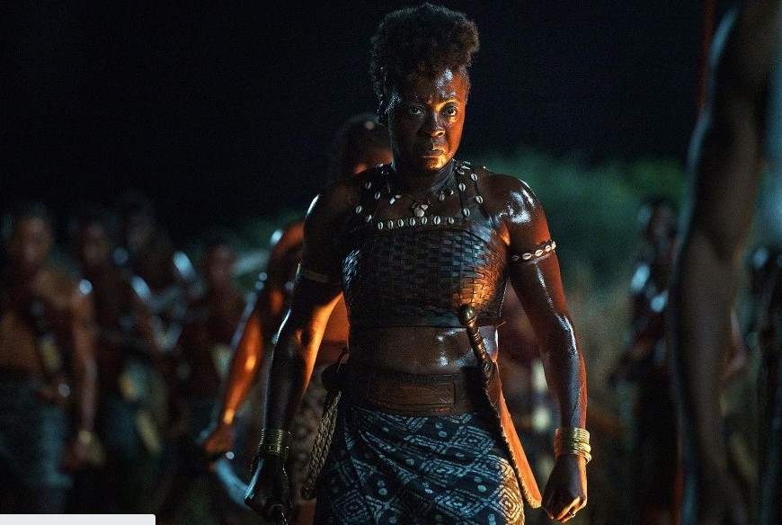 A black woman in her fifties stands on a battlefield at night in African dress. She wears a sword at her waist and a fierce look.